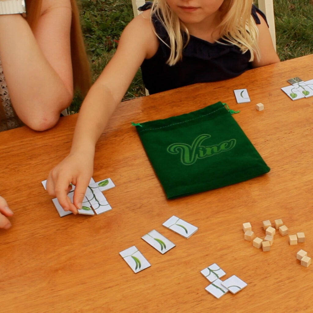 Adult and child playing Vine at an outdoor table with tiles in play, beans in the market area, and growth tokens on the table